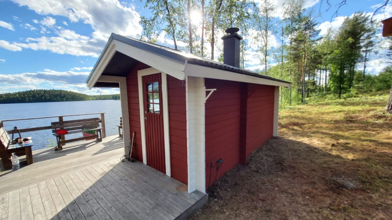 klassisches rotes schwedisches Saunahaus am See mit Sonnenterrasse bei strahlendem Sonnenschein in Abborrträsk, Norrbotten.