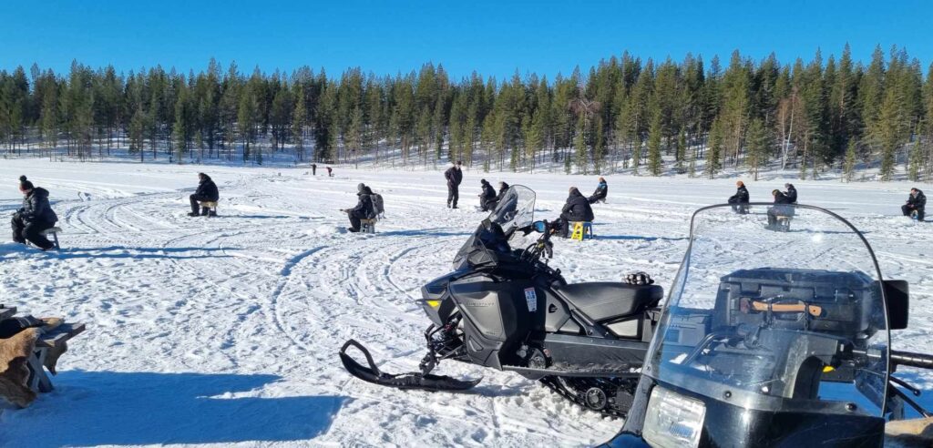 Viele Personen sitzen auf dem gefrorenen See bei Sonnenschein und angeln.